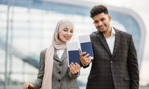 Smiling Muslim woman in hijab and Arab man, posing on camera outside modern airport terminal, demonstrating their passports and tickets while waiting flight. Ready for trip. Business people